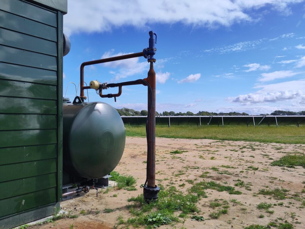 Ende eines kleinen grünen Tanks hinter gründer Gebäudeecke vor Reihe von Sonnenkollektoren am Horizont.