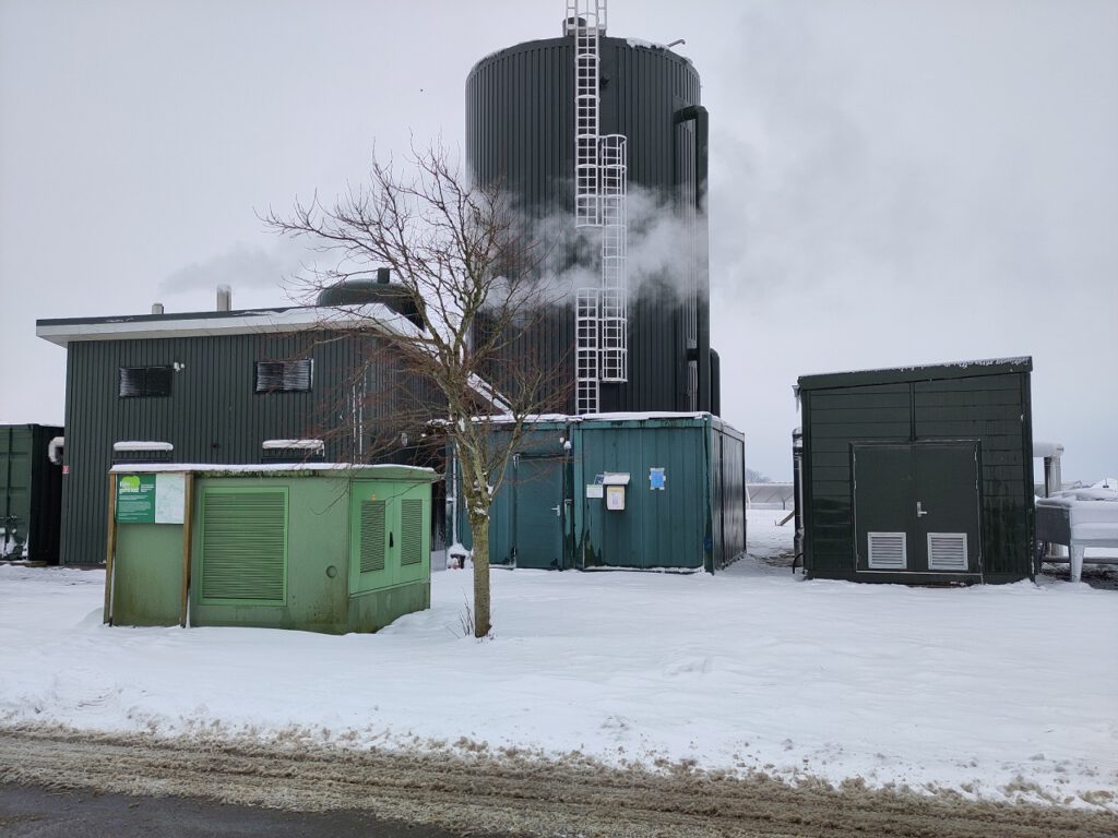 Hinter den Gebäuden der Heizzentrale sind der kleine und der große Wärmespeicher zu sehen. Ganz klein ist auch ein Kollektor der Solarthermieanlage zu erkennen. Vorne auf eine Schneefläche ein kleiner kahler Baum neben einem Trafoblock.
